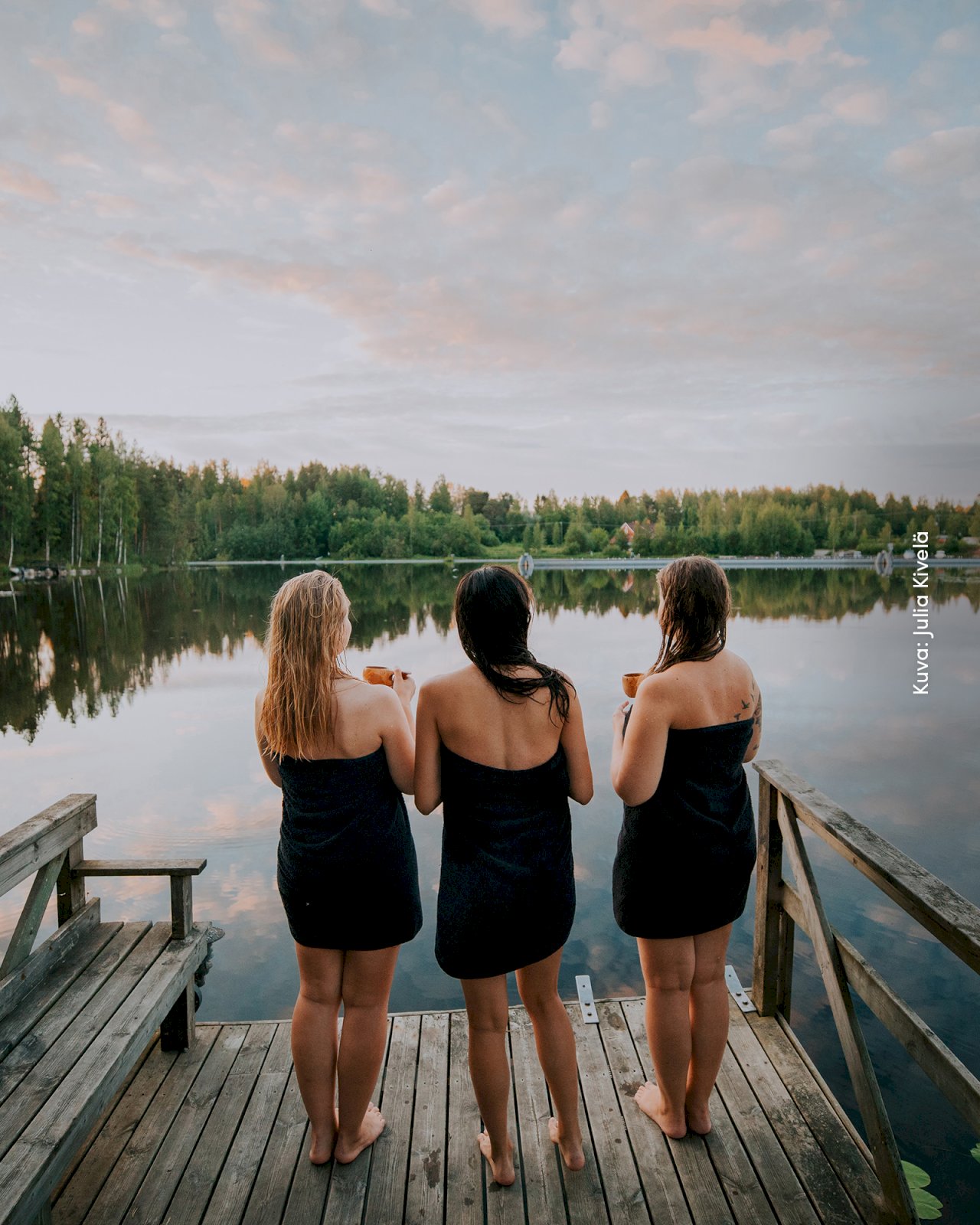 Kolme naista seisovat pyyhkeet päällä Varjolan laiturilla katsellen järvimaisemaa auringonlaskussa.